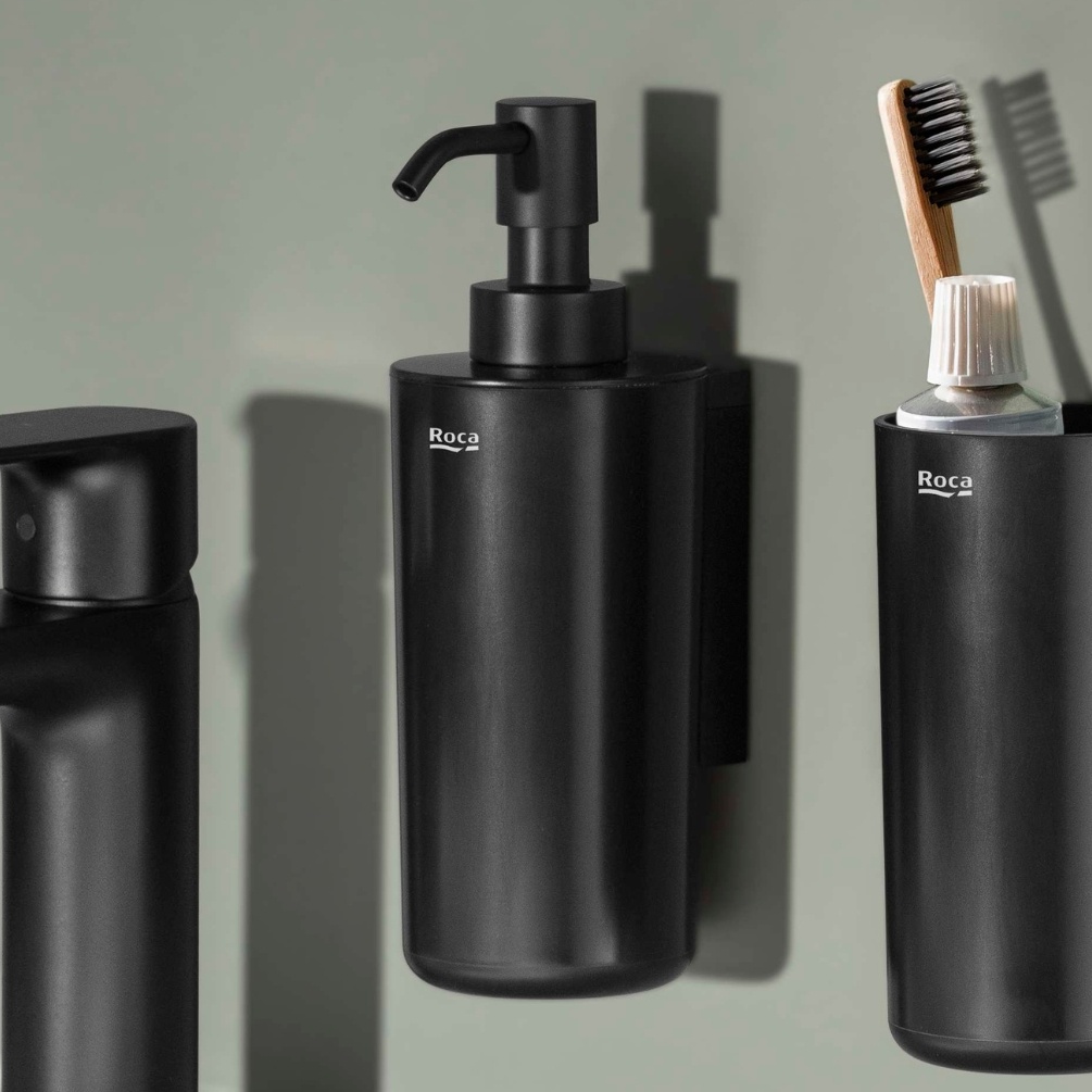 Lifestyle image of the Roca Sonata Matt Black Wall-Mounted Soap Dispenser mounted on a wall above a basin and next to a toothbrush holder